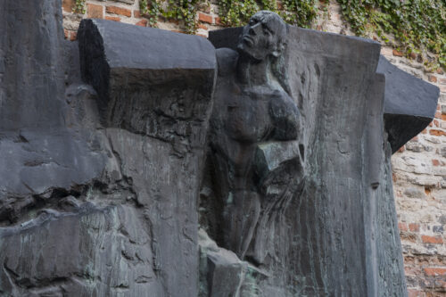 Zdjęcie ukazuje rzeźbę z brązu z patyną, na której można zauważyć postacie ludzkie wyłaniające się z form przypominających mur lub gruzy. Rzeźba została wkomponowana w ścianę z cegieł.