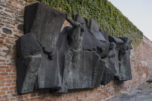 Zdjęcie ukazuje rzeźbę z brązu z patyną, na której można zauważyć postacie ludzkie wyłaniające się z form przypominających mur lub gruzy. Rzeźba została wkomponowana w ścianę z cegieł.