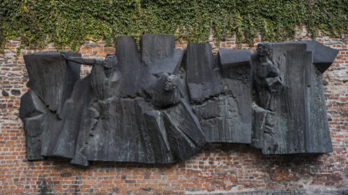Zdjęcie ukazuje rzeźbę z brązu z patyną, na której można zauważyć postacie ludzkie wyłaniające się z form przypominających mur lub gruzy. Rzeźba została wkomponowana w ścianę z cegieł.