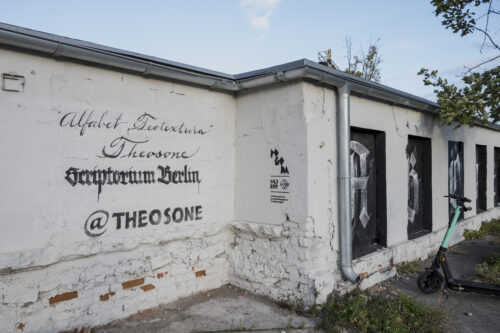 Zdjęcie przedstawia niski budynek pomalowany na biało, którego okna zasłonięto czarną dyktą, na których namalowano litery alfabetu krojem pisma przypominającego gotyckie