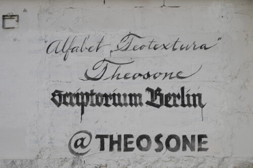 Zdjęcie pokazuje fragment muru pomalowanego na biało, na którym widać czarne napisy - podpis do pracy.