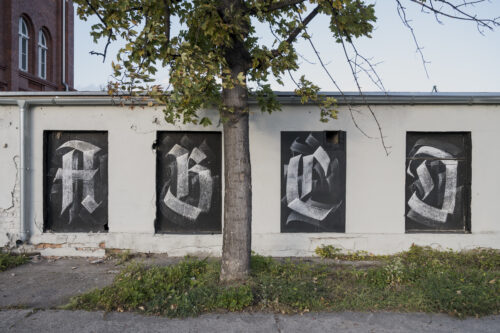 Zdjęcie przedstawia niski budynek pomalowany na biało, którego okna zasłonięto czarną dyktą, na których namalowano litery alfabetu krojem pisma przypominającego gotyckie