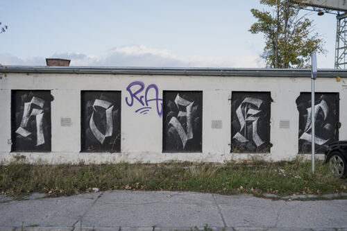 Zdjęcie przedstawia niski budynek pomalowany na biało, którego okna zasłonięto czarną dyktą, na których namalowano litery alfabetu krojem pisma przypominającego gotyckie