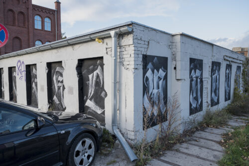 Zdjęcie przedstawia niski budynek pomalowany na biało, którego okna zasłonięto czarną dyktą, na których namalowano litery alfabetu krojem pisma przypominającego gotyckie