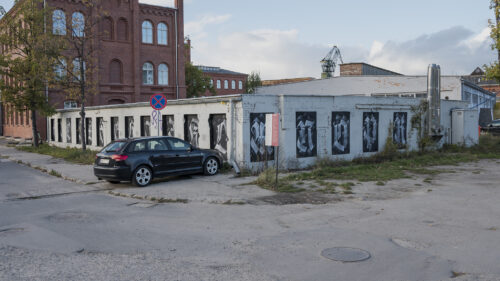 Zdjęcie przedstawia niski budynek pomalowany na biało, którego okna zasłonięto czarną dyktą, na których namalowano litery alfabetu krojem pisma przypominającego gotyckie.