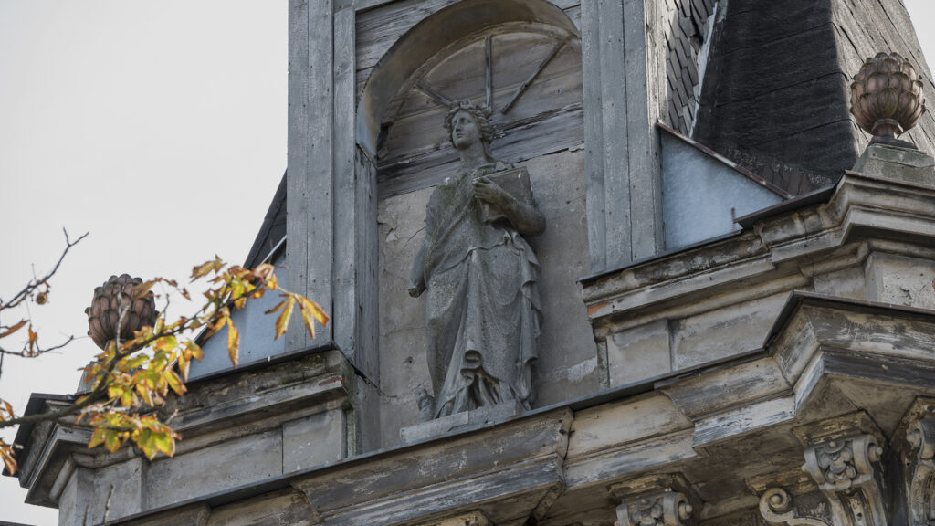 Zdjęcie poziome ze zbliżeniem na szczyt jednej z zabytkowych kamienic w przestrzeni miejskiej. Znajduje się tam koncha, w której wnętrzu umieszczono pełnopostaciową figurę kobiety. Postać odziana jest w starożytną togę, w swojej lewej dłoni trzyma książkę, zaś na głowie dostrzegalny jest roślinny wieniec.