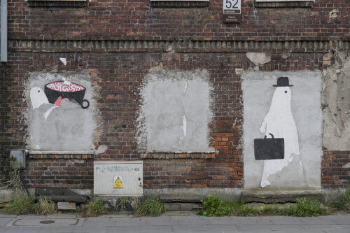 Zdjęcie ukazuje fragment budynku z czerwonej cegły, w którego oknach wstawiono płyty na których naniesiono rysunki duchów na białym tle.