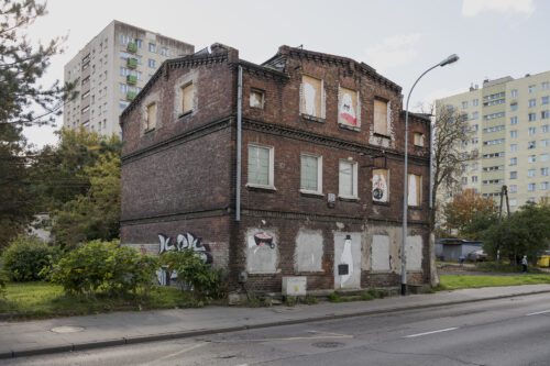 Zdjęcie poziomie ukazujące z ukosa opuszczony budynek mieszkalny z czerwonej cegły z zamurowanyim oknami, w których umieszczono rysunki duchów.