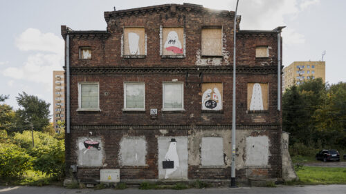 Poziome zdjęcie ukazujące przedwojenny budynek z czerwonej cegły z zabitymi dyktą oknami i zamurowanym wejściem. W niektórych oknach umieszczono rysunki przedstawiające rysunkowe duchy.