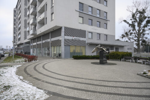 Zdjęcie poziomie przedstawiające przestrzeń miejską z zabudową mieszkalną. Przed wejście do bloku została ustawiona metalowa rzeźba na ciemnym, geometrycznym postumencie.