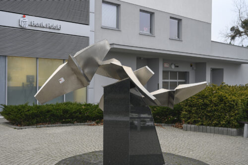 Zdjęcie poziomie przedstawiające geometryczną, abstrakcyjną rzeźbę wykonaną z metalu, która została ustawiona na ciemnym postumencie. Figura swoim kształtem ptaka w trakcie lotu. W tle znajduje się nowoczesny blok mieszkalny.