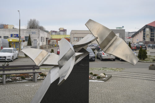 Zdjęcie poziomie przedstawiające geometryczną, abstrakcyjną rzeźbę wykonaną z metalu, która została ustawiona na ciemnym postumencie. Figura swoim kształtem ptaka w trakcie lotu. W tle znajdują się niskie pawilony handlowe.