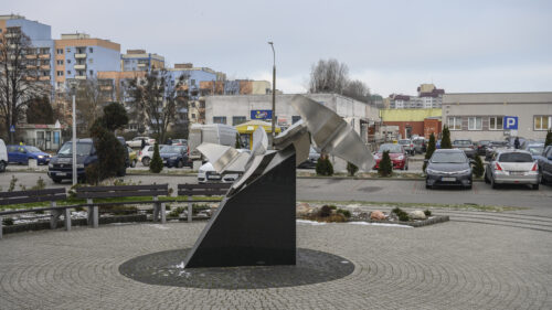 Zdjęcie poziomie przedstawiające geometryczną, abstrakcyjną rzeźbę wykonaną z metalu, która została ustawiona na ciemnym postumencie. Figura swoim kształtem ptaka w trakcie lotu. W tle znajdują się niskie pawilony handlowe oraz wielopiętrowe bloki mieszkalne.