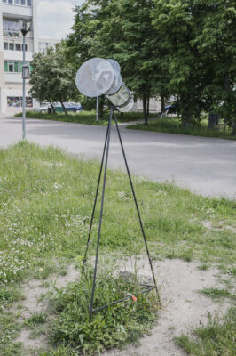 Zdjęcie pionowe przedstawiające instalację artystyczną w przestrzeni osiedla bloków mieszkaniowych. Instalacja swoją konstrukcją przypomina przezroczystą lunetę obserwacyjną na wysokim, cienkim statywie. Skierowana jest ona ku górze, jednak na jej obiektyw zostały nałożone trzy transparentne koła. Instalacja została umieszczona bezpośrednio na trawniku.