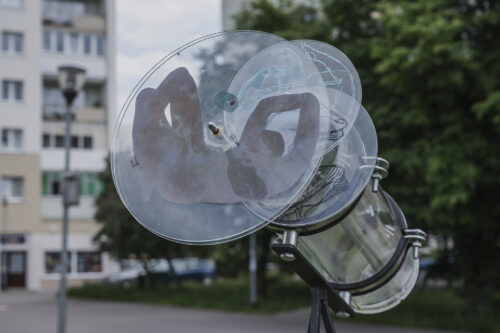 Zdjęcie poziome przedstawiające instalację artystyczną w przestrzeni osiedla bloków mieszkaniowych. Instalacja swoją konstrukcją przypomina skierowaną ku górze, przezroczystą lunetę obserwacyjną. Na jej obiektyw zostały nałożone trzy transparentne koła, których powierzchnie pokryto plamami barwnymi i rysunkami.