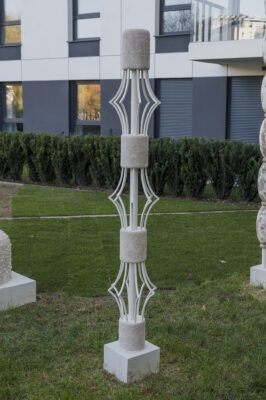 Wertykalna forma rzeźbiarska w białym kolorze przypominająca totem, złożona z kilku modułów o geometrycznych, abstrakcyjnych kształtach ułożonych na sobie. Co drugi moduł posiada ażurowe prześwity.
