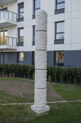 Wertykalna rzeźba w kolorze białym o chropowatej fakturze przypomina kolumnę o kształcie walca złożonego z modułów. W tle zabudowa osiedlowa.