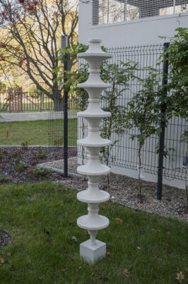 Wertykalna rzeźba w kolorze białym z fakturą, przypomina totem złożóny z geometrycznych walcowatych dysków. Rzeźba stoi na trawie, w tle zieleń i budynki.