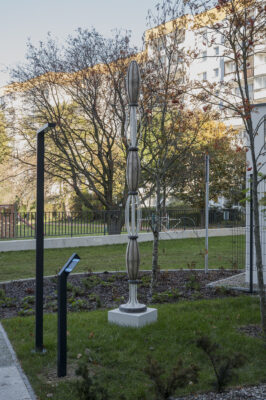Ażurowa, pionowa rzeźba złożona z wrzecionowatych geometrycznych kształtów ułożonych jeden na drugim, w tle zieleń miejska i zabudowa osiedla.